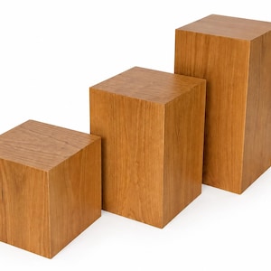 Könnte beinhalten: Drei Holz-Ausstellungsblöcke unterschiedlicher Höhe. Die Blöcke sind quadratisch und haben eine natürliche Holzmaserung. Die Blöcke sind in einer abgestuften Konfiguration angeordnet, wodurch eine visuelle Hierarchie entsteht. Ideal für die Präsentation von Produkten.