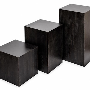 May include: Three dark brown wooden display cubes of varying heights. The cubes are arranged in a stepped configuration, with the smallest cube on the left and the tallest on the right. The wood grain is visible.