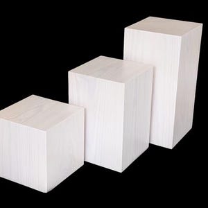 Può includere: Tre cubi espositivi in legno bianco di altezze variabili, disposti a gradini. I cubi hanno una texture di venatura del legno chiaro e sono su uno sfondo nero, ideali per esporre prodotti.