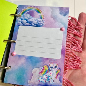 Puede incluir: Un peque&ntilde;o cuaderno abierto con anillas doradas y una cubierta verde ne&oacute;n. La p&aacute;gina interior presenta una colorida ilustraci&oacute;n de un gato con alas de arco&iacute;ris, un arco&iacute;ris y nubes. Una borla rosa est&aacute; unida al lateral.