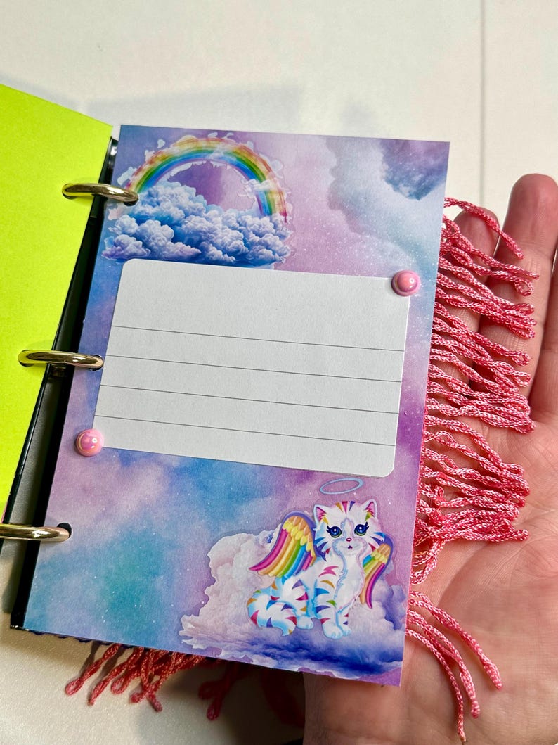 Puede incluir: Un peque&ntilde;o cuaderno abierto con anillas doradas y una cubierta verde ne&oacute;n. La p&aacute;gina interior presenta una colorida ilustraci&oacute;n de un gato con alas de arco&iacute;ris, un arco&iacute;ris y nubes. Una borla rosa est&aacute; unida al lateral.