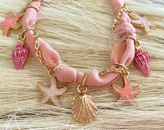 Op de afbeelding: Een roze stoffen armband met gouden kettingaccenten en roze en witte schelpbedels. De bedels omvatten zeesterren, schelpen en een schelpbedel.