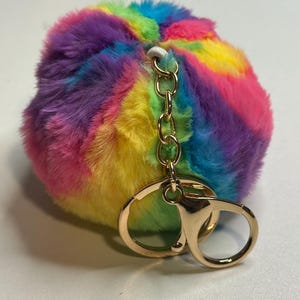 Puede incluir: Un llavero de pomp&oacute;n multicolor. El llavero presenta un cierre y una cadena de color dorado. El pomp&oacute;n es una mezcla de colores vibrantes, incluyendo rosa, amarillo, verde, azul y morado, creando un accesorio l&uacute;dico y llamativo.