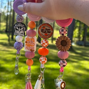 Puede incluir: Una colección de carretes de insignia con cuentas en tonos rosa, naranja y morado. Cada carrete presenta cuentas decorativas, dijes y un cordón retráctil con un clip. Los dijes incluyen un girasol, una calabaza y el texto "Take Me To The Pumpkin Patch".