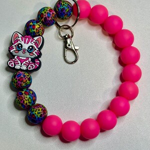 Puede incluir: Una pulsera con cuentas rosas para llavero con un dije de gato de dibujos animados. La pulsera presenta una mezcla de cuentas rosas brillantes y coloridas con estampado de leopardo. Se adjunta un cierre y un llavero plateados.