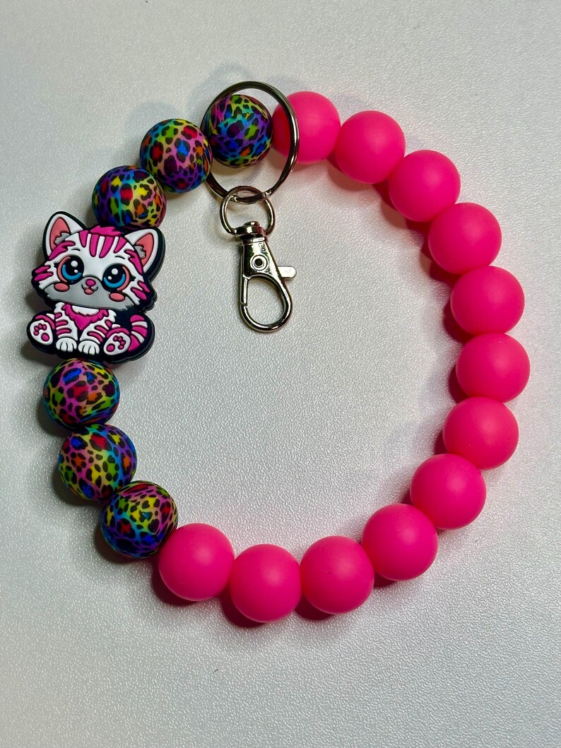 Puede incluir: Una pulsera con cuentas rosas para llavero con un dije de gato de dibujos animados. La pulsera presenta una mezcla de cuentas rosas brillantes y coloridas con estampado de leopardo. Se adjunta un cierre y un llavero plateados.