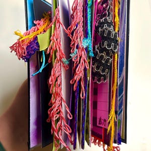 Puede incluir: Un diario hecho a mano con p&aacute;ginas coloridas y elementos decorativos. El diario presenta borlas rojas, rosas y amarillas, junto con detalles de encaje azul y negro. Las p&aacute;ginas son una mezcla de colores, incluyendo morado, rosa y azul.