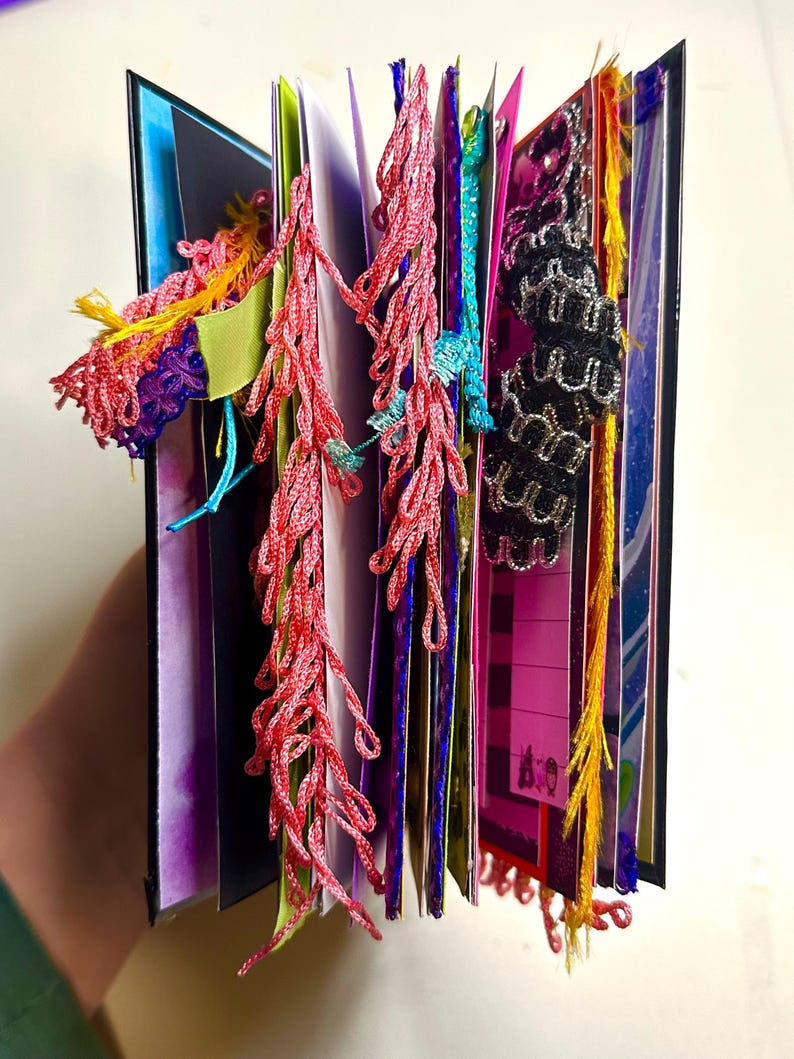 Puede incluir: Un diario hecho a mano con p&aacute;ginas coloridas y elementos decorativos. El diario presenta borlas rojas, rosas y amarillas, junto con detalles de encaje azul y negro. Las p&aacute;ginas son una mezcla de colores, incluyendo morado, rosa y azul.