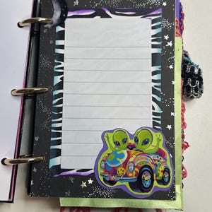 Puede incluir: Un cuaderno con espiral con una p&aacute;gina rayada. El cuaderno presenta una cubierta negra con un patr&oacute;n de rayas de cebra, estrellas y una luna creciente. Una pegatina colorida de dos extraterrestres verdes en un coche psicod&eacute;lico est&aacute; en la p&aacute;gina.