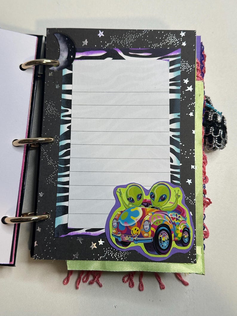 Puede incluir: Un cuaderno con espiral con una p&aacute;gina rayada. El cuaderno presenta una cubierta negra con un patr&oacute;n de rayas de cebra, estrellas y una luna creciente. Una pegatina colorida de dos extraterrestres verdes en un coche psicod&eacute;lico est&aacute; en la p&aacute;gina.