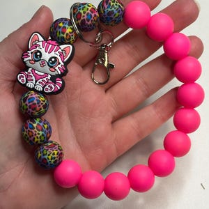 Puede incluir: Una pulsera de cuentas rosa ne&oacute;n con un cierre plateado y un dije con un gato de dibujos animados con rayas rosas y blancas. La pulsera tambi&eacute;n incluye cuentas con un estampado de leopardo colorido.