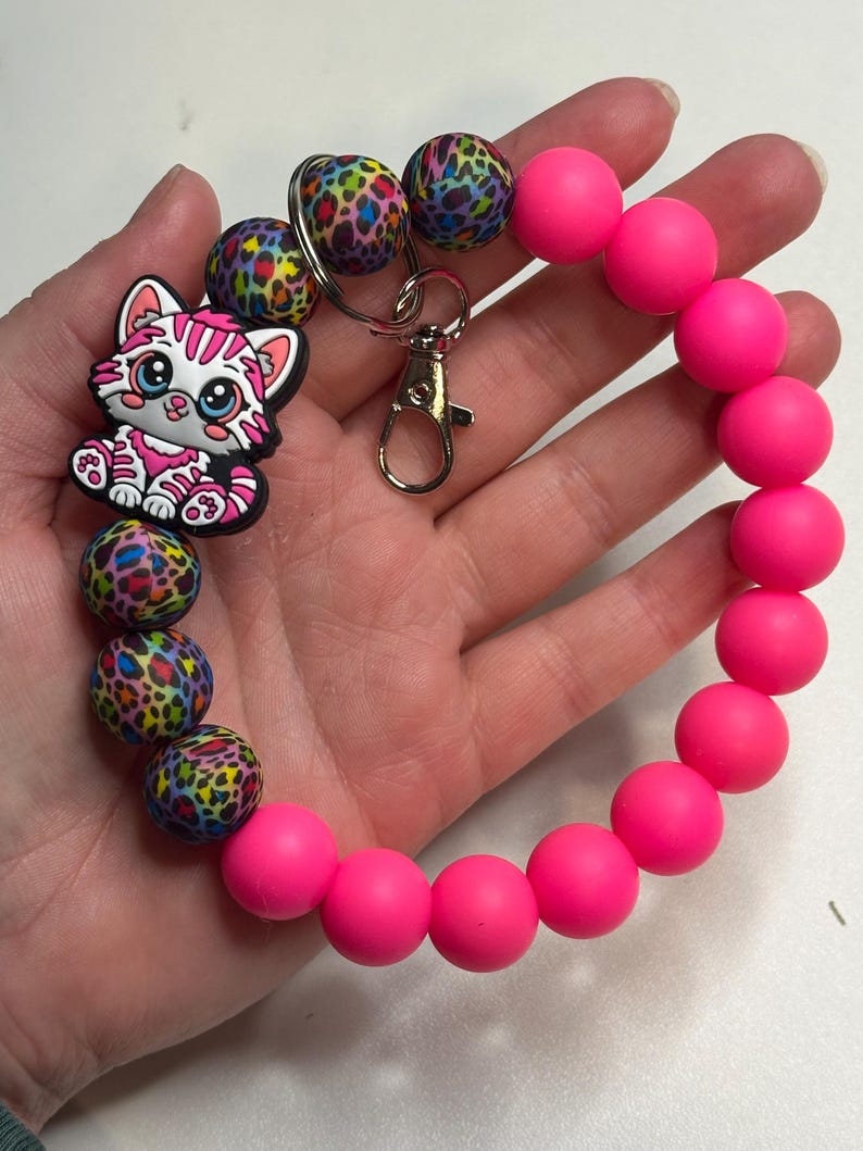 Puede incluir: Una pulsera de cuentas rosa ne&oacute;n con un cierre plateado y un dije con un gato de dibujos animados con rayas rosas y blancas. La pulsera tambi&eacute;n incluye cuentas con un estampado de leopardo colorido.