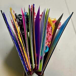 Puede incluir: Un libro abierto y colorido con p&aacute;ginas desplegadas, sujetas por un anillo dorado. Las p&aacute;ginas son de varios colores, incluyendo rosa, morado y verde, con borlas y hilos decorativos.