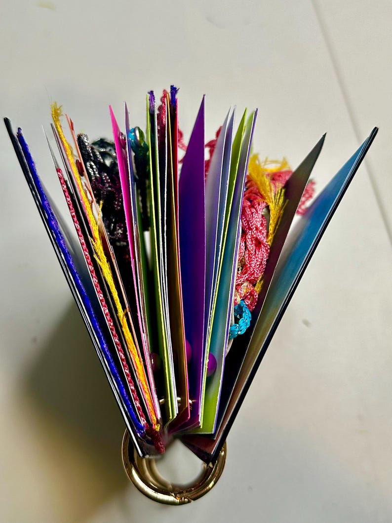 Puede incluir: Un libro abierto y colorido con p&aacute;ginas desplegadas, sujetas por un anillo dorado. Las p&aacute;ginas son de varios colores, incluyendo rosa, morado y verde, con borlas y hilos decorativos.