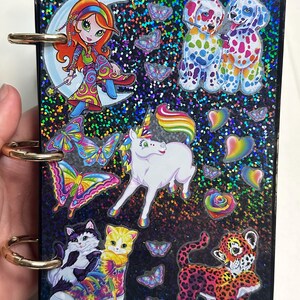 Puede incluir: Un peque&ntilde;o cuaderno negro brillante con anillos dorados. La portada presenta pegatinas de dibujos animados coloridos de una ni&ntilde;a en una luna creciente, dos perros manchados, un unicornio, mariposas, gatos y un leopardo.