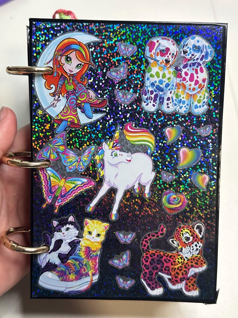 Puede incluir: Un peque&ntilde;o cuaderno negro brillante con anillos dorados. La portada presenta pegatinas de dibujos animados coloridos de una ni&ntilde;a en una luna creciente, dos perros manchados, un unicornio, mariposas, gatos y un leopardo.