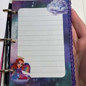 Puede incluir: Una p&aacute;gina de cuaderno con l&iacute;neas y una colorida ilustraci&oacute;n de un &aacute;ngel con alas de arco&iacute;ris y un gato azul. La p&aacute;gina tiene un fondo de cielo estrellado y una pegatina de nube. El cuaderno est&aacute; encuadernado con anillas met&aacute;licas y tiene un ribete de encaje morado.