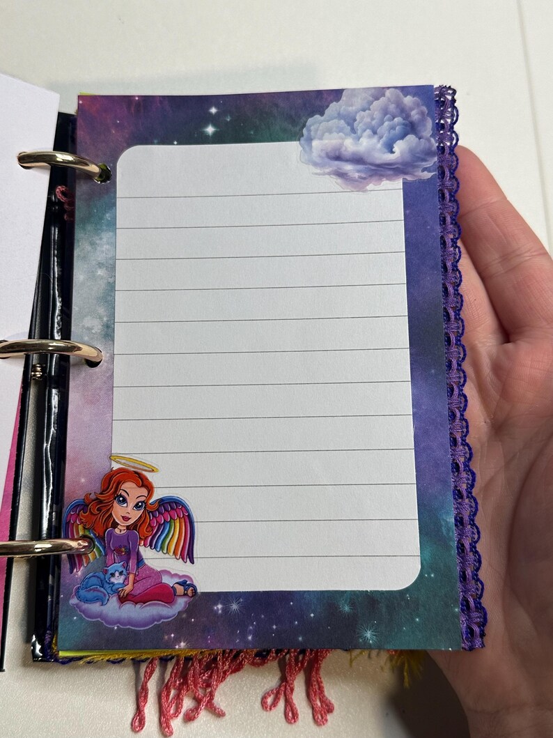 Puede incluir: Una p&aacute;gina de cuaderno con l&iacute;neas y una colorida ilustraci&oacute;n de un &aacute;ngel con alas de arco&iacute;ris y un gato azul. La p&aacute;gina tiene un fondo de cielo estrellado y una pegatina de nube. El cuaderno est&aacute; encuadernado con anillas met&aacute;licas y tiene un ribete de encaje morado.