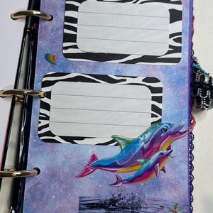 Puede incluir: Una p&aacute;gina de diario colorida con un fondo de galaxia, marcos de etiquetas con estampado de cebra y un gr&aacute;fico de delfines arco&iacute;ris. El diario est&aacute; encuadernado con anillas met&aacute;licas y tiene un borde morado con borlas rosas.