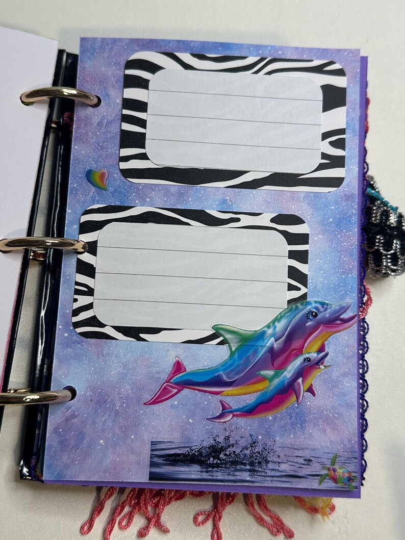 Puede incluir: Una p&aacute;gina de diario colorida con un fondo de galaxia, marcos de etiquetas con estampado de cebra y un gr&aacute;fico de delfines arco&iacute;ris. El diario est&aacute; encuadernado con anillas met&aacute;licas y tiene un borde morado con borlas rosas.