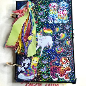 Puede incluir: Un diario colorido con una cubierta negra, adornado con pegatinas de un unicornio, perros, gatos y un leopardo. El diario presenta un arco&iacute;ris de colores, incluyendo mariposas, corazones y una zapatilla deportiva te&ntilde;ida. Una borla decorativa cuelga de la encuadernaci&oacute;n del diario.