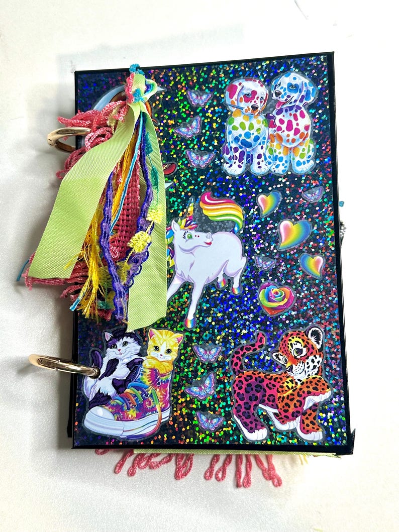 Puede incluir: Un diario colorido con una cubierta negra, adornado con pegatinas de un unicornio, perros, gatos y un leopardo. El diario presenta un arco&iacute;ris de colores, incluyendo mariposas, corazones y una zapatilla deportiva te&ntilde;ida. Una borla decorativa cuelga de la encuadernaci&oacute;n del diario.