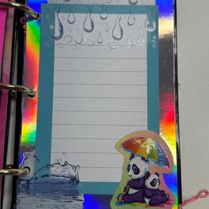 Puede incluir: Una p&aacute;gina de scrapbook colorida con un papel azul rayado y un borde hologr&aacute;fico. La p&aacute;gina presenta una pegatina de dos pandas bajo un paraguas arco&iacute;ris. Los gr&aacute;ficos de gotas de lluvia y salpicaduras de agua se suman al tema.