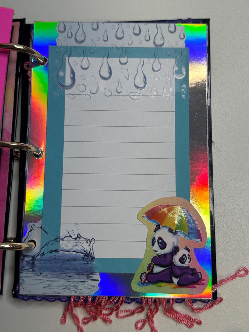 Puede incluir: Una p&aacute;gina de scrapbook colorida con un papel azul rayado y un borde hologr&aacute;fico. La p&aacute;gina presenta una pegatina de dos pandas bajo un paraguas arco&iacute;ris. Los gr&aacute;ficos de gotas de lluvia y salpicaduras de agua se suman al tema.