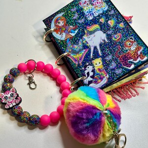 Puede incluir: Un juego de llaveros coloridos con una pulsera de cuentas rosas con un dije de gato, una bola de peluche de arco&iacute;ris y un peque&ntilde;o cuaderno con pegatinas de animales y criaturas fant&aacute;sticas. El cuaderno tiene anillos dorados.