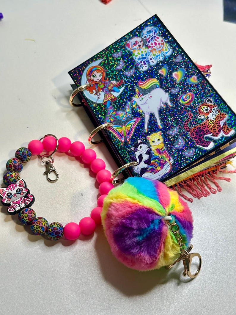 Puede incluir: Un juego de llaveros coloridos con una pulsera de cuentas rosas con un dije de gato, una bola de peluche de arco&iacute;ris y un peque&ntilde;o cuaderno con pegatinas de animales y criaturas fant&aacute;sticas. El cuaderno tiene anillos dorados.