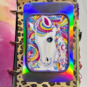 Puede incluir: Un diario con una imagen de unicornio sobre un fondo hologr&aacute;fico. El unicornio tiene una cara blanca y una melena arco&iacute;ris. La imagen est&aacute; enmarcada con un borde de cuerda morada. El diario tiene una cubierta con estampado de leopardo.