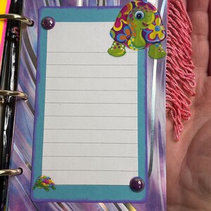 Puede incluir: Una p&aacute;gina de cuaderno colorida con un borde turquesa y papel rayado. Una pegatina de tortuga de dibujos animados con un dise&ntilde;o floral est&aacute; en la parte superior, y una pegatina de tortuga m&aacute;s peque&ntilde;a en la parte inferior. La p&aacute;gina est&aacute; decorada con detalles morados y flecos rosas.