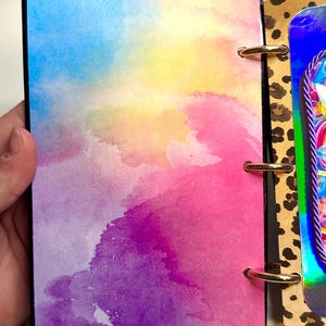 Puede incluir: Un diario colorido con un dise&ntilde;o de acuarela. La portada presenta una mezcla de tonos azules, amarillos, rosas y morados. El diario est&aacute; encuadernado con anillos dorados y tiene un borde negro. La p&aacute;gina adyacente tiene un dise&ntilde;o de estampado de leopardo.