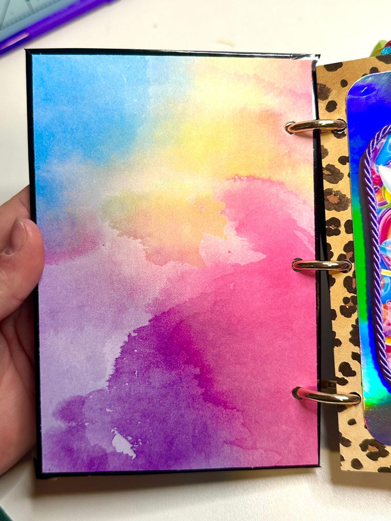 Puede incluir: Un diario colorido con un dise&ntilde;o de acuarela. La portada presenta una mezcla de tonos azules, amarillos, rosas y morados. El diario est&aacute; encuadernado con anillos dorados y tiene un borde negro. La p&aacute;gina adyacente tiene un dise&ntilde;o de estampado de leopardo.