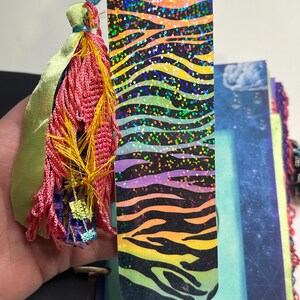 Puede incluir: Un marcap&aacute;ginas colorido con un dise&ntilde;o de rayas de cebra en tonos naranja, amarillo, morado y verde, con detalles de purpurina. Tiene una borla hecha de hilos rosas, amarillos y azules, y una cinta verde.