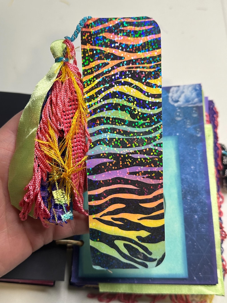 Puede incluir: Un marcap&aacute;ginas colorido con un dise&ntilde;o de rayas de cebra en tonos naranja, amarillo, morado y verde, con detalles de purpurina. Tiene una borla hecha de hilos rosas, amarillos y azules, y una cinta verde.