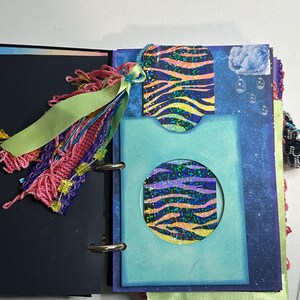 Puede incluir: Diario abierto con una cubierta azul y negra. En el interior, una tarjeta azul con un recorte circular revela un dise&ntilde;o estampado. Una etiqueta con el mismo dise&ntilde;o cuelga de la parte superior. Una borla y una cinta coloridas adornan el lomo del diario.