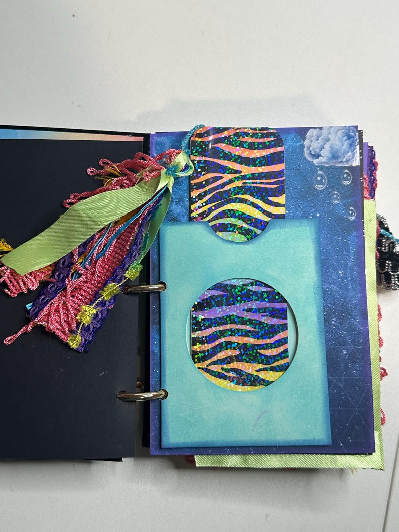 Puede incluir: Diario abierto con una cubierta azul y negra. En el interior, una tarjeta azul con un recorte circular revela un dise&ntilde;o estampado. Una etiqueta con el mismo dise&ntilde;o cuelga de la parte superior. Una borla y una cinta coloridas adornan el lomo del diario.