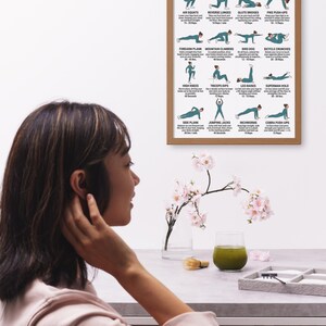 May include: A framed poster titled "Military Calisthenics Workout for Women" displays illustrated exercise instructions. The poster is in a light brown frame. A woman is seen in the foreground, with a table holding a glass of green liquid and a small floral arrangement.