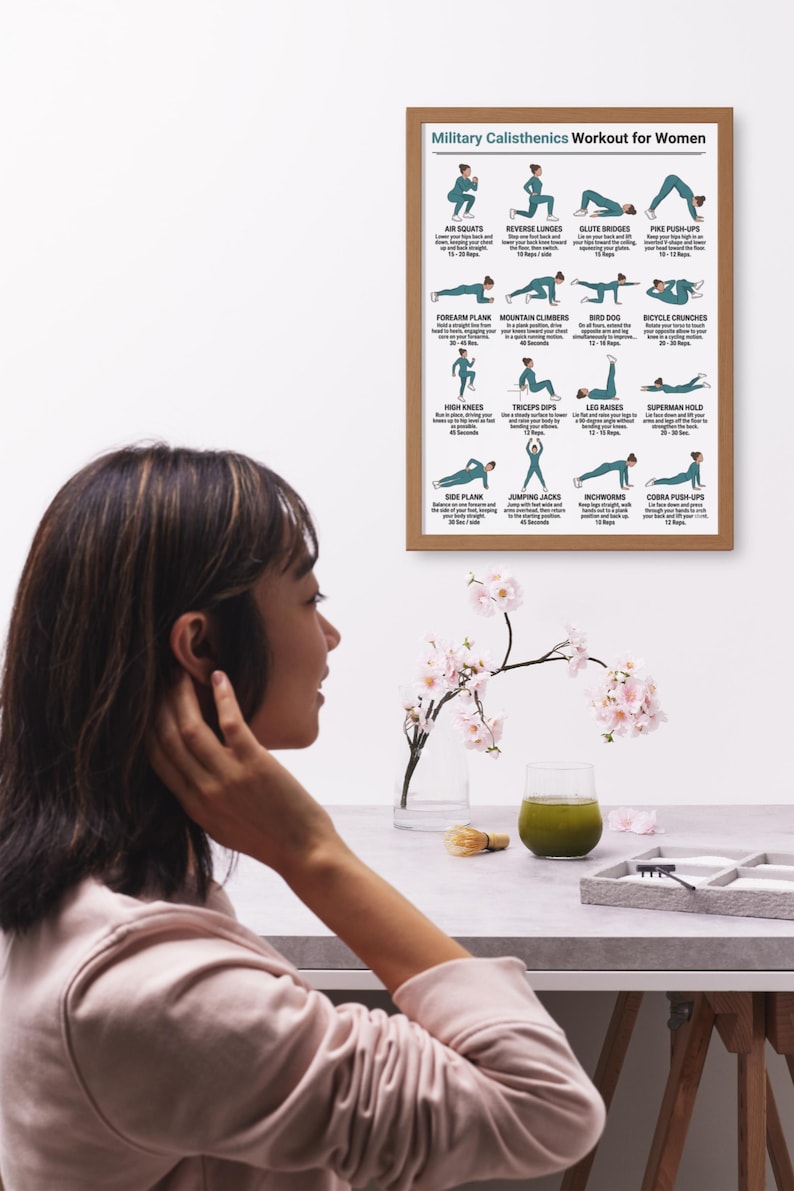 May include: A framed poster titled "Military Calisthenics Workout for Women" displays illustrated exercise instructions. The poster is in a light brown frame. A woman is seen in the foreground, with a table holding a glass of green liquid and a small floral arrangement.