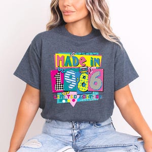 May include: Dark gray t-shirt with a colorful graphic that reads "MADE IN 1986 LIMITED EDITION." The graphic features the numbers 1, 9, and 6 in a variety of patterns and colors, including pink, yellow, and black and white checkerboard.