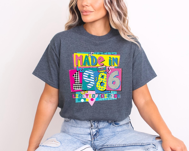 May include: Dark gray t-shirt with a colorful graphic that reads "MADE IN 1986 LIMITED EDITION." The graphic features the numbers 1, 9, and 6 in a variety of patterns and colors, including pink, yellow, and black and white checkerboard.