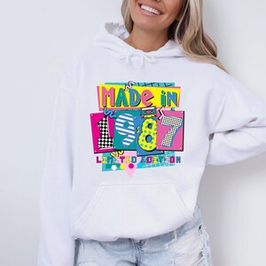 Może przedstawiać: Biała bluza z kapturem z kolorową grafiką z przodu. Grafika głosi "MADE IN 1987 LIMITED EDITION" w stylu retro. Projekt zawiera cyfry 1, 9, 8 i 7 w r&oacute;żnych wzorach i kolorach.
