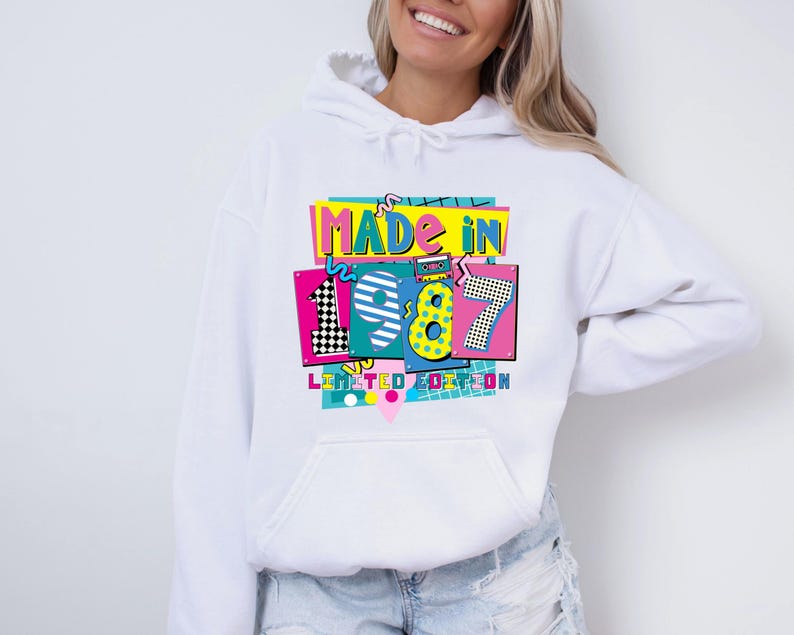 Może przedstawiać: Biała bluza z kapturem z kolorową grafiką z przodu. Grafika głosi "MADE IN 1987 LIMITED EDITION" w stylu retro. Projekt zawiera cyfry 1, 9, 8 i 7 w r&oacute;żnych wzorach i kolorach.