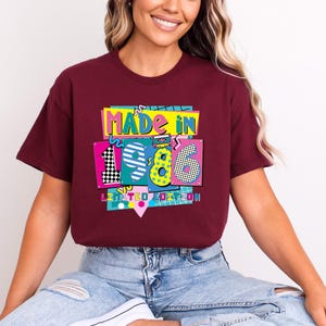 May include: A maroon t-shirt with a colorful graphic that reads "MADE IN 1986 LIMITED EDITION." The graphic features the numbers 1, 9, and 6 in different patterns and colors, with a cassette tape design. The shirt is worn by a person.