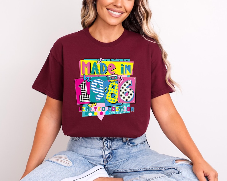 May include: A maroon t-shirt with a colorful graphic that reads "MADE IN 1986 LIMITED EDITION." The graphic features the numbers 1, 9, and 6 in different patterns and colors, with a cassette tape design. The shirt is worn by a person.