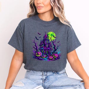 May include: A dark heather gray short-sleeve t-shirt featuring a vibrant graphic print of a haunted house, bare trees, flying bats, and a bright green moon. Three glowing purple and pink jack-o'-lanterns with carved faces sit in the foreground.