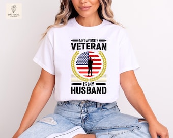 May include: White t-shirt with the text "MY FAVORITE VETERAN IS MY HUSBAND" in black. The design includes an American flag and a silhouette of a soldier within a gold wreath. The shirt is a classic crew neck style.