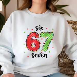 May include: White sweatshirt with a colorful graphic featuring the numbers "6" and "7" decorated with lights. The number "6" is red with white polka dots, and the number "7" is green with white polka dots. The word "six" and "seven" are written above and below the numbers.