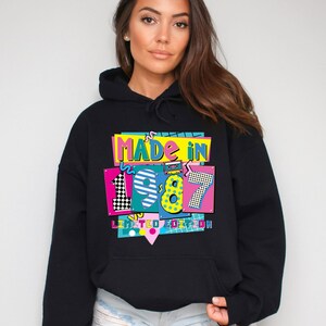 Może przedstawiać: Czarna bluza z kapturem z kolorową grafiką z napisem "MADE IN 1987 LIMITED EDITION". Grafika ma retro design z żywymi kolorami, w tym r&oacute;żowym, ż&oacute;łtym, niebieskim i zielonym. Bluza ma kaptur ze sznurkiem.